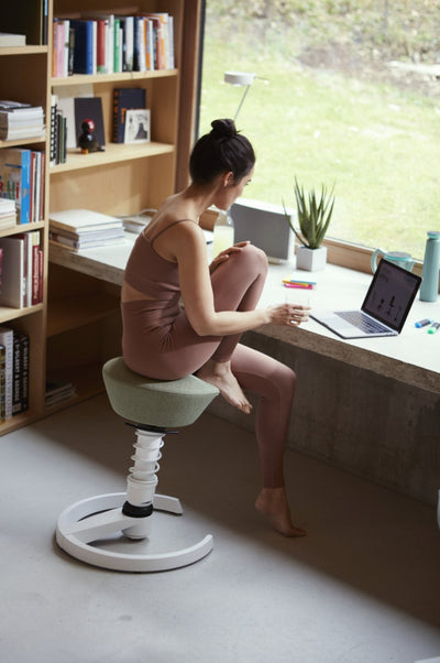 A woman sitting on an Aeris Swopper which is an ergonomic office with an iconic spring a convex formed polstered seat. She is looking at her laptop in front of a big window